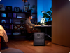 BLUETTI Elite 320 Portable Power Station with 1,800 W Output | 3,200 Wh , LCD display and multiple ports on a wooden floor with a man sat at a desk working on a computer with a blue background with shelves www.mygeneratorplus.com 