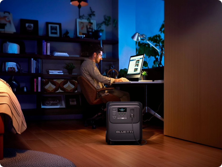 BLUETTI Elite 320 Portable Power Station with 1,800 W Output | 3,200 Wh , LCD display and multiple ports on a wooden floor with a man sat at a desk working on a computer with a blue background with shelves www.mygeneratorplus.com 